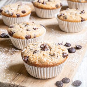 Lightly dusted chocolate chip muffins on a wooden board surrounded by lots of chocolate chips.