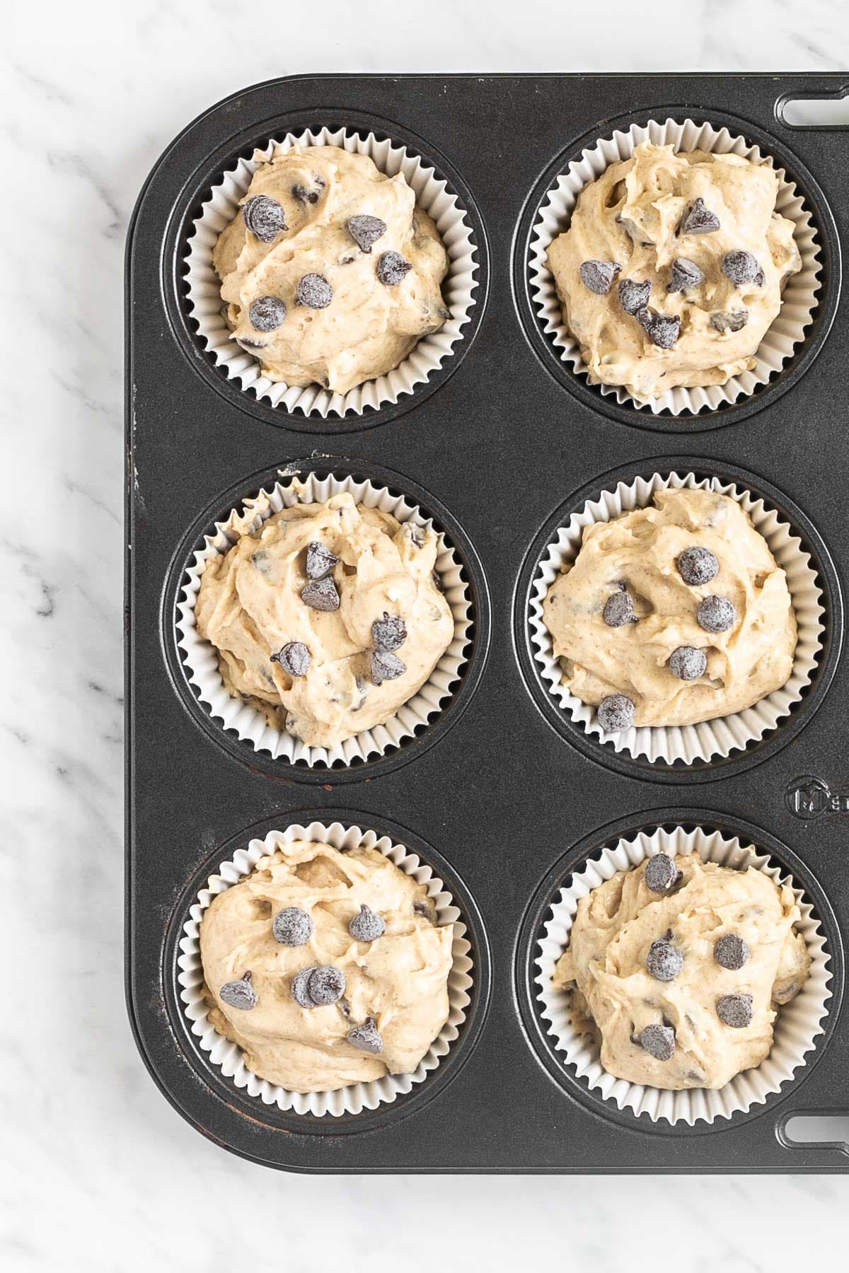A muffin tin lined with white cupcake liners and filled with light brown batter with chocolate chips to the top.
