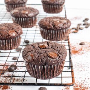 Several chocolate muffins with chocolate chips on a black wire rack. Even more chocolate chips are scattered around and under the rack.