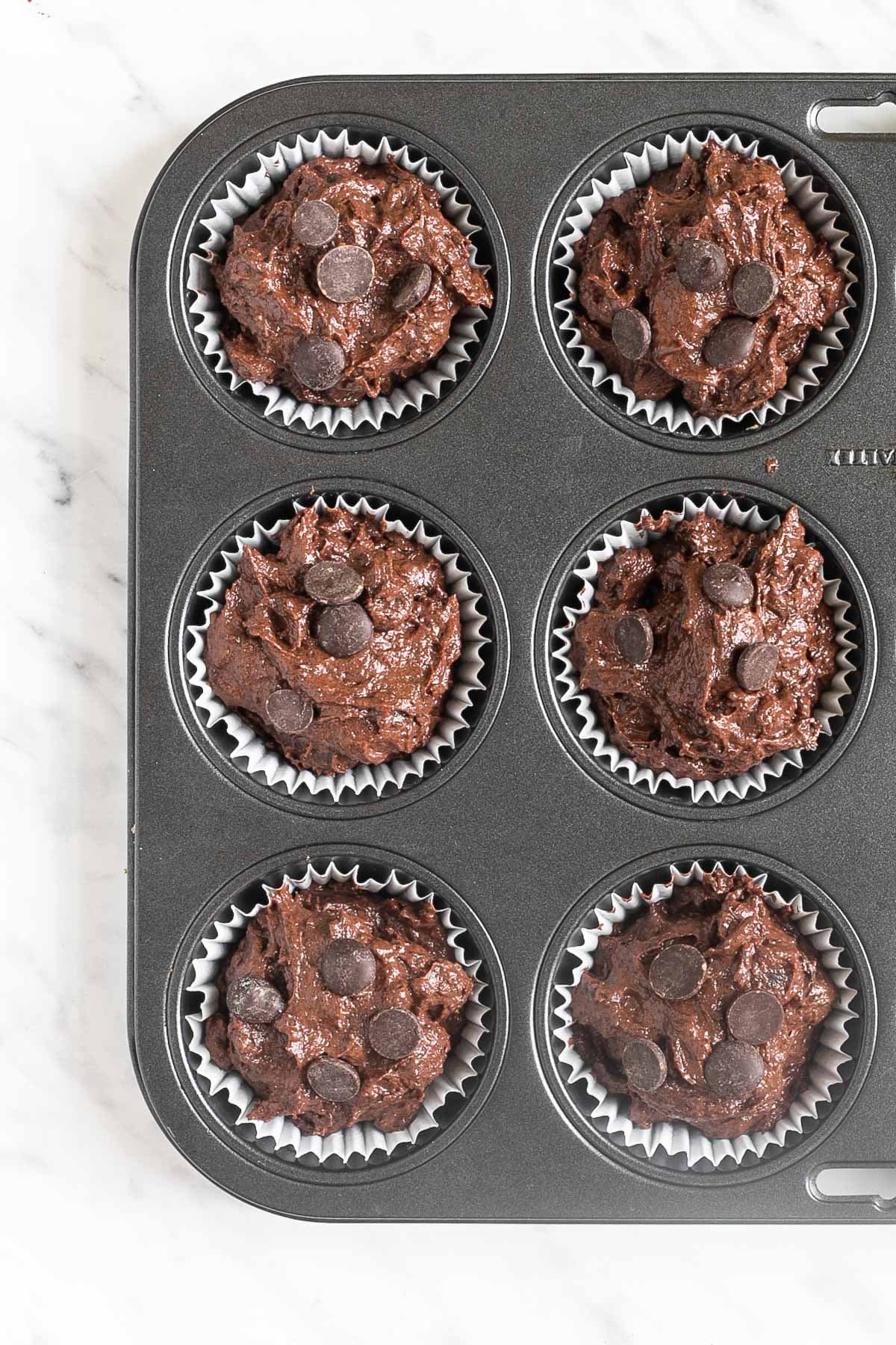 A muffin tin lined with white cupcake liners and filled with dark brown batter mixed with chocolate chips