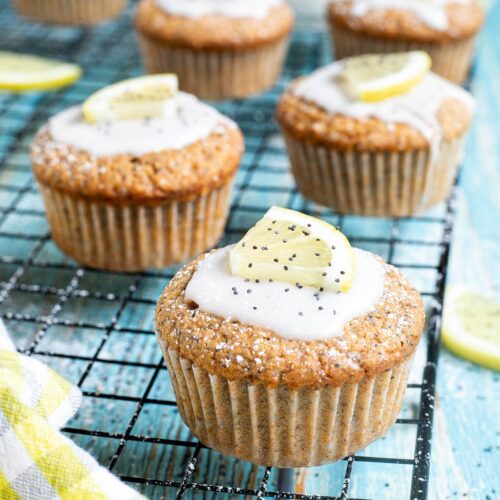 Several lemon poppy seed muffins on a black wire rack topped with a white glaze and a small lemon slices, and sprinkled with poppy seeds. Lemon slices are scattered around them.