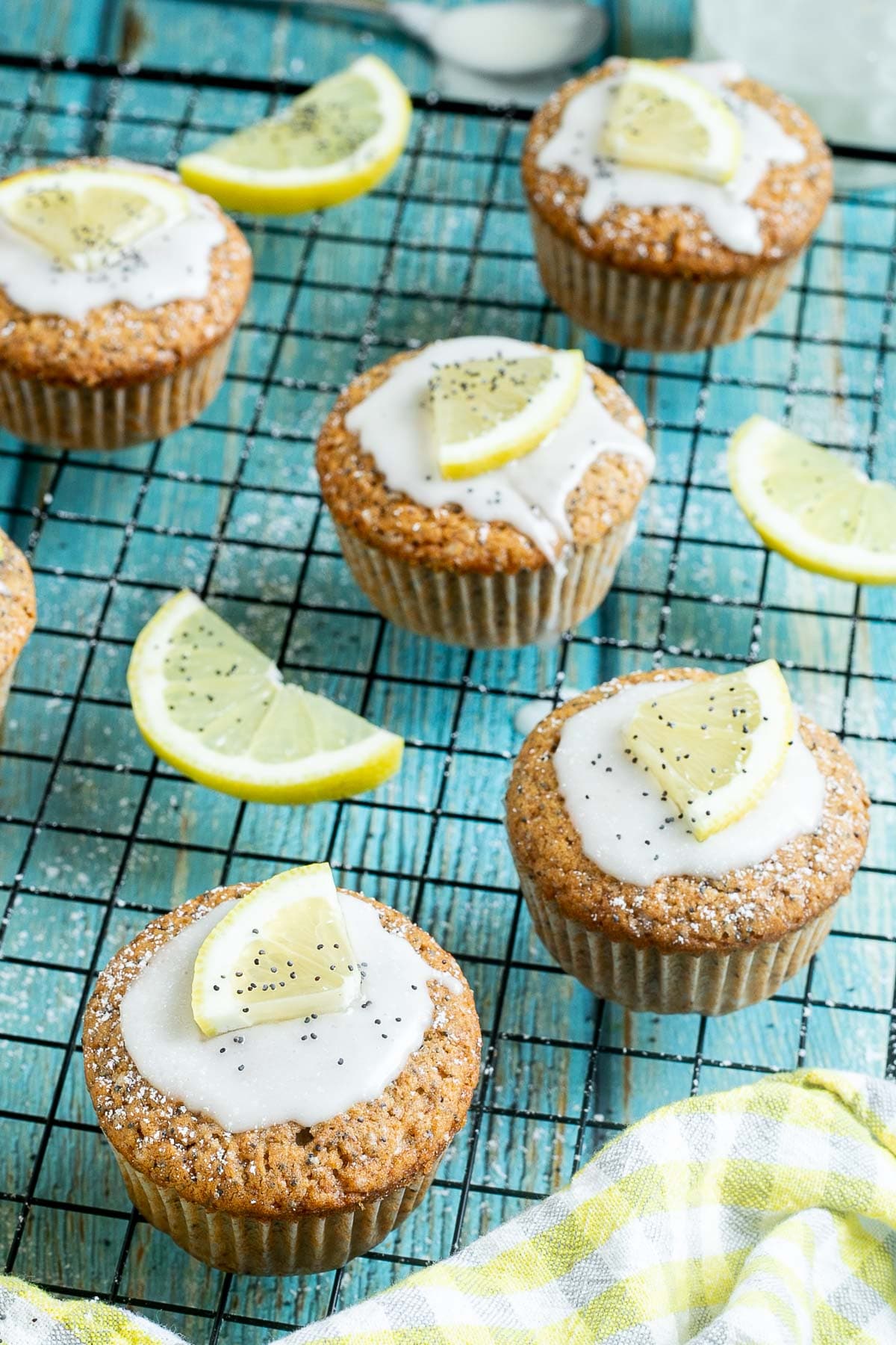 Several lemon poppy seed muffins on a black wire rack topped with a white glaze and a small lemon slices, and sprinkled with poppy seeds. Lemon slices are scattered around them.