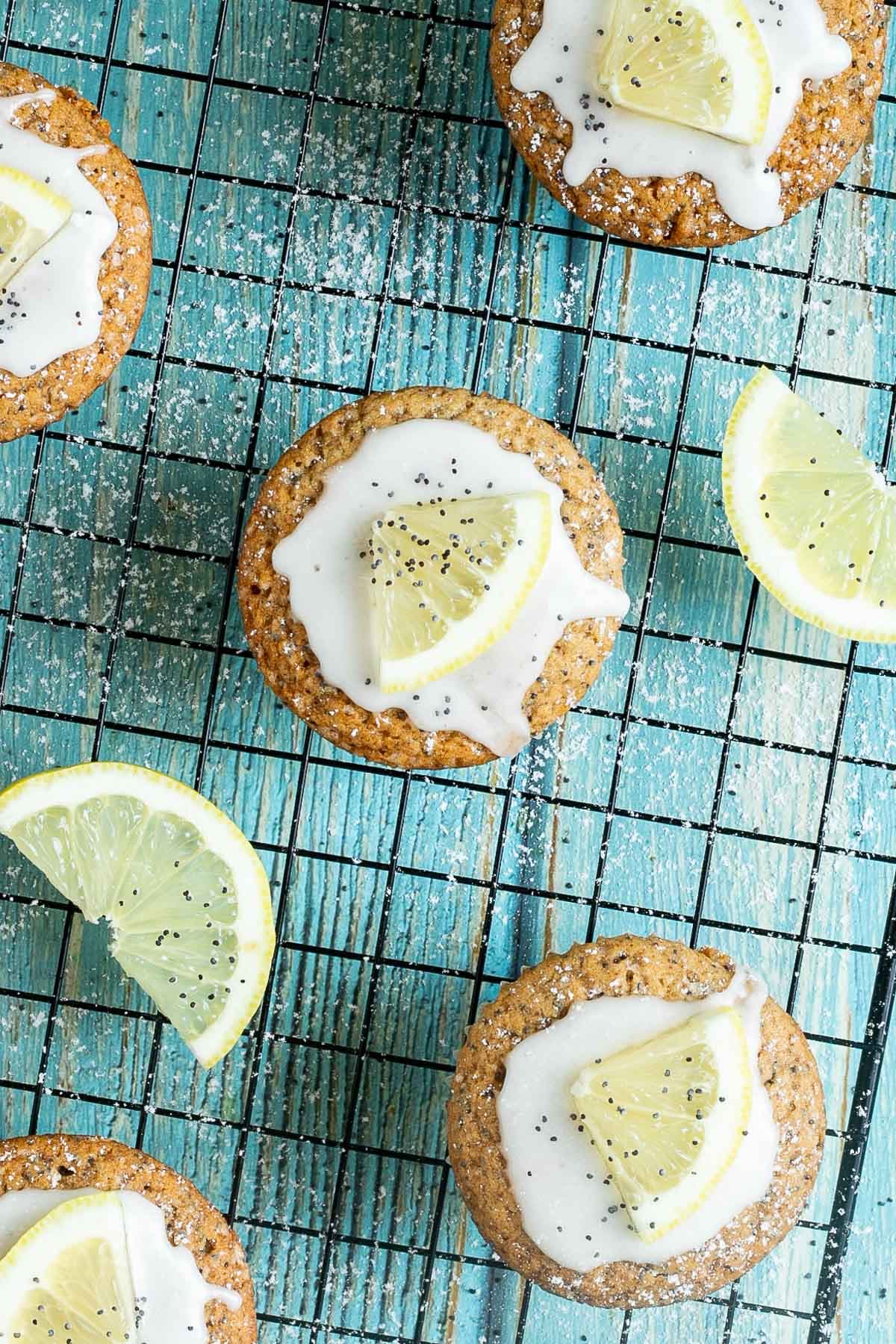 Several lemon poppy seed muffins on a black wire rack topped with a white glaze and a small lemon slices, and sprinkled with poppy seeds. Lemon slices are scattered around them.