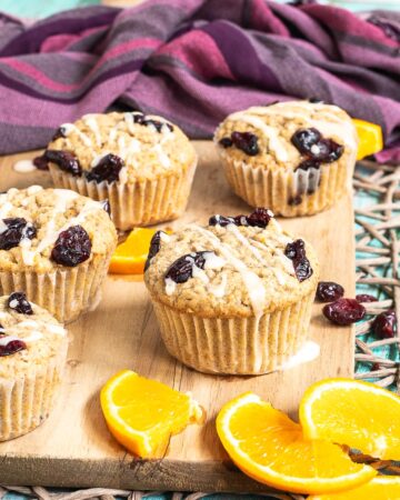 Orange cranberry muffins drizzled with a white glaze on a wooden board surrounded by lots of dried cranberries and fresh orange slices.
