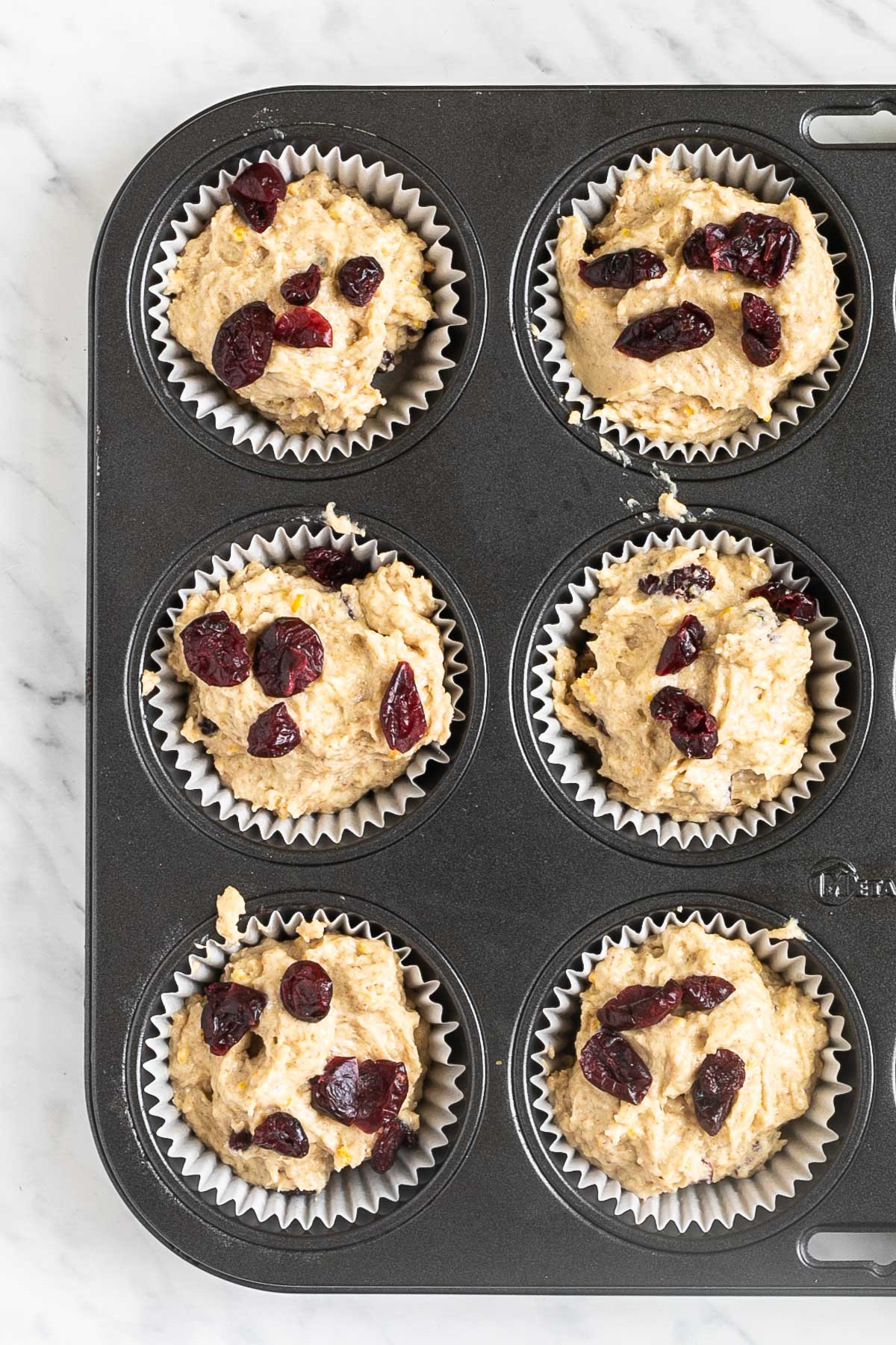 A muffin tin lined with white cupcake liners and filled with light brown batter with dried cranberries to the top.