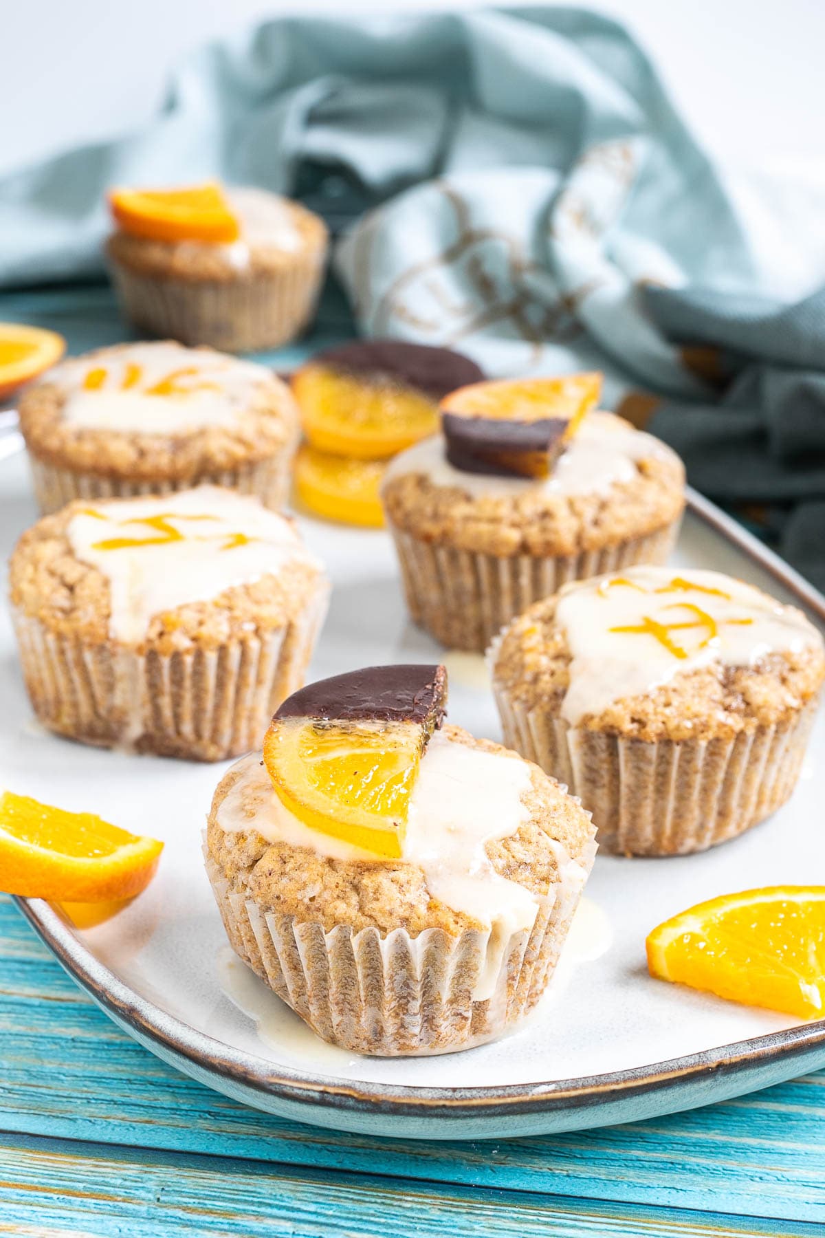 Several orange muffins on a light blue plate topped with a white glaze and orange slices partly dipped in chocolate or white glaze with orange zest pieces. Orange slices are scattered around them.