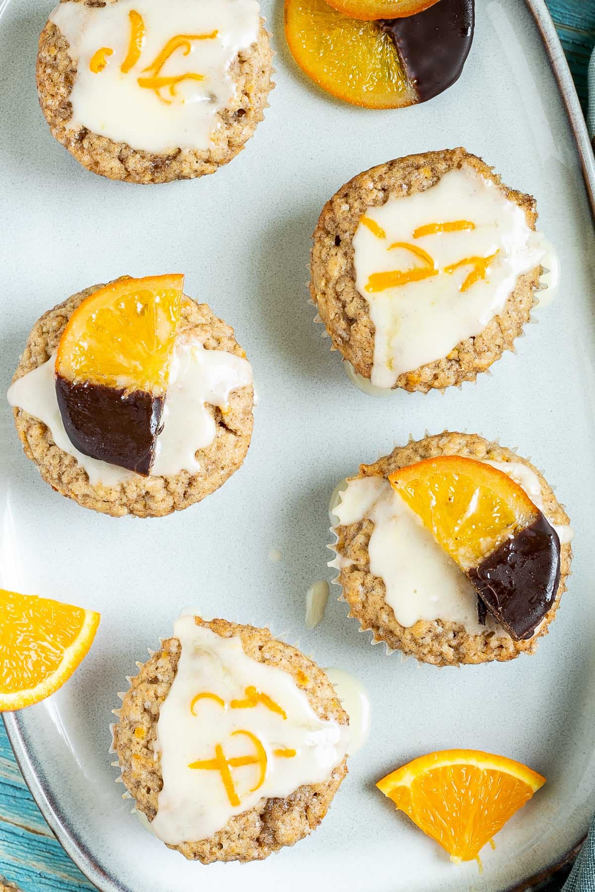 Several orange muffins on a light blue plate topped with a white glaze and orange slices partly dipped in chocolate or white glaze with orange zest pieces. Orange slices are scattered around them.