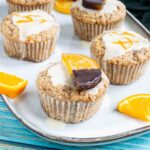 Several orange muffins on a light blue plate topped with a white glaze and orange slices partly dipped in chocolate or white glaze with orange zest pieces. Orange slices are scattered around them.