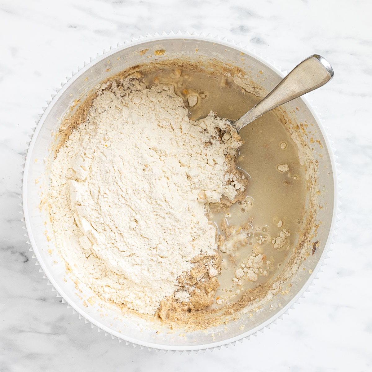 Liquid and a heap of flour is on top of a soft light brown batter in a white bowl.