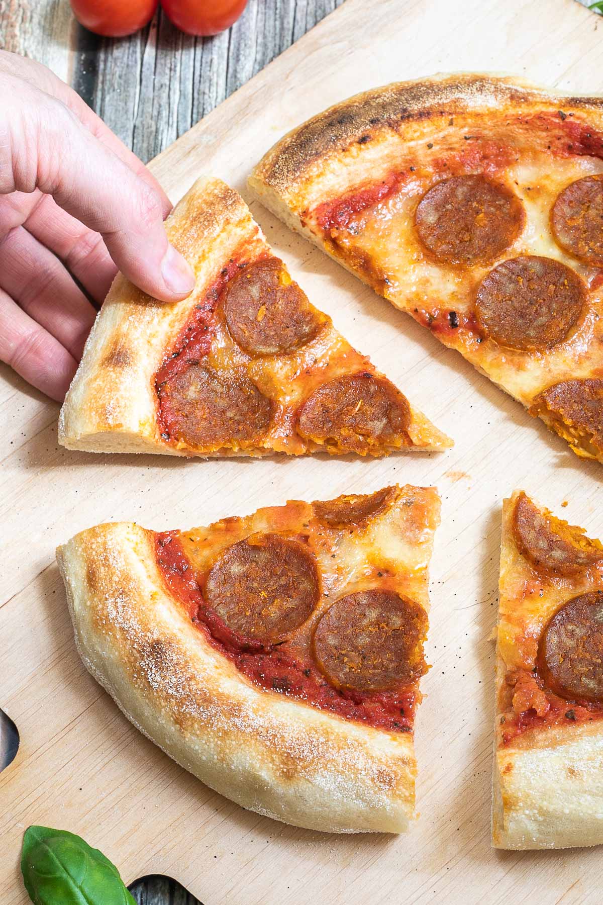 Sliced pizza on a wooden baker's peel topped with tomato sauce, melted cheese and pepperoni slices. A hand is taking a slice.