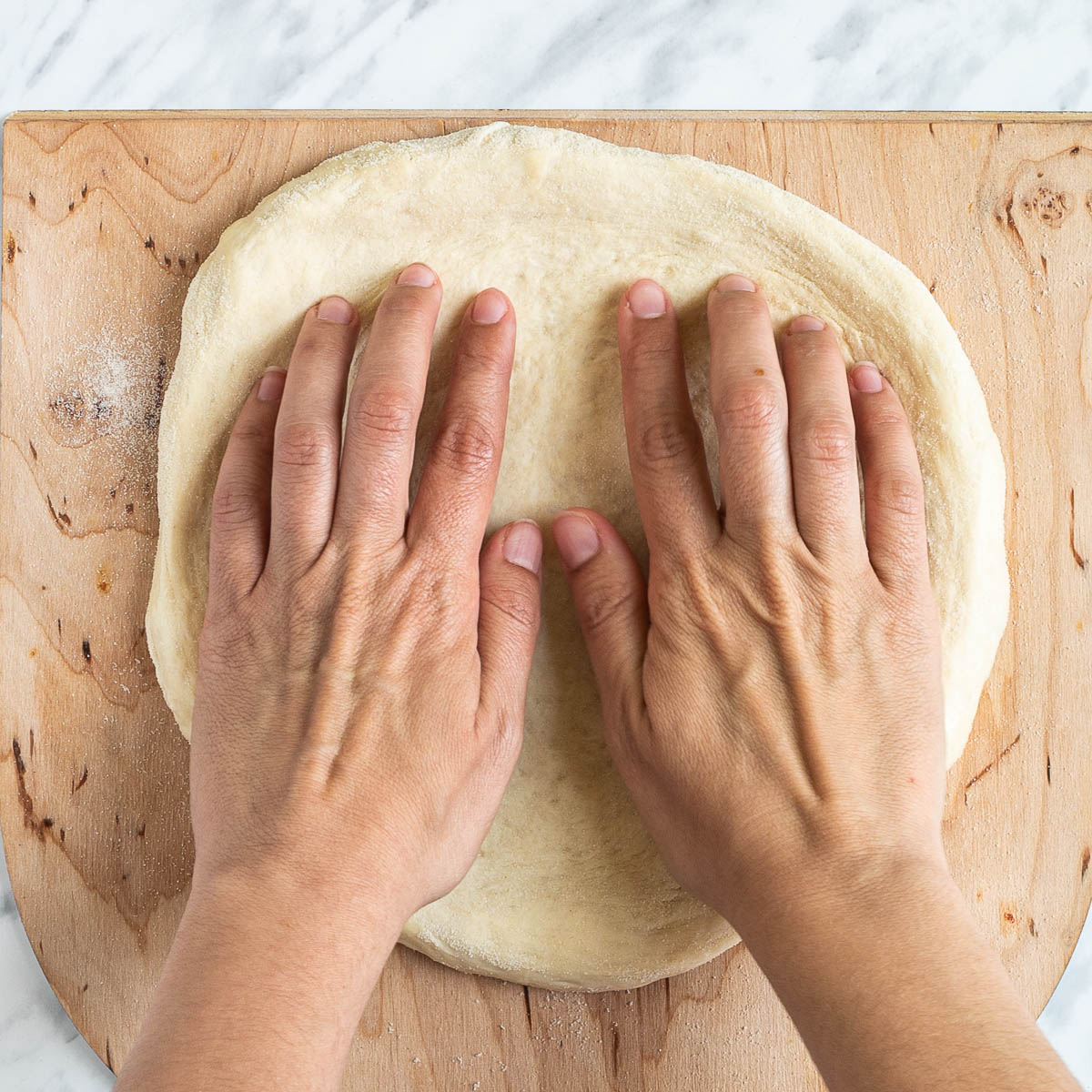 Two hands are shaping the pizza dough on a lightly floured wooden pizza peel.