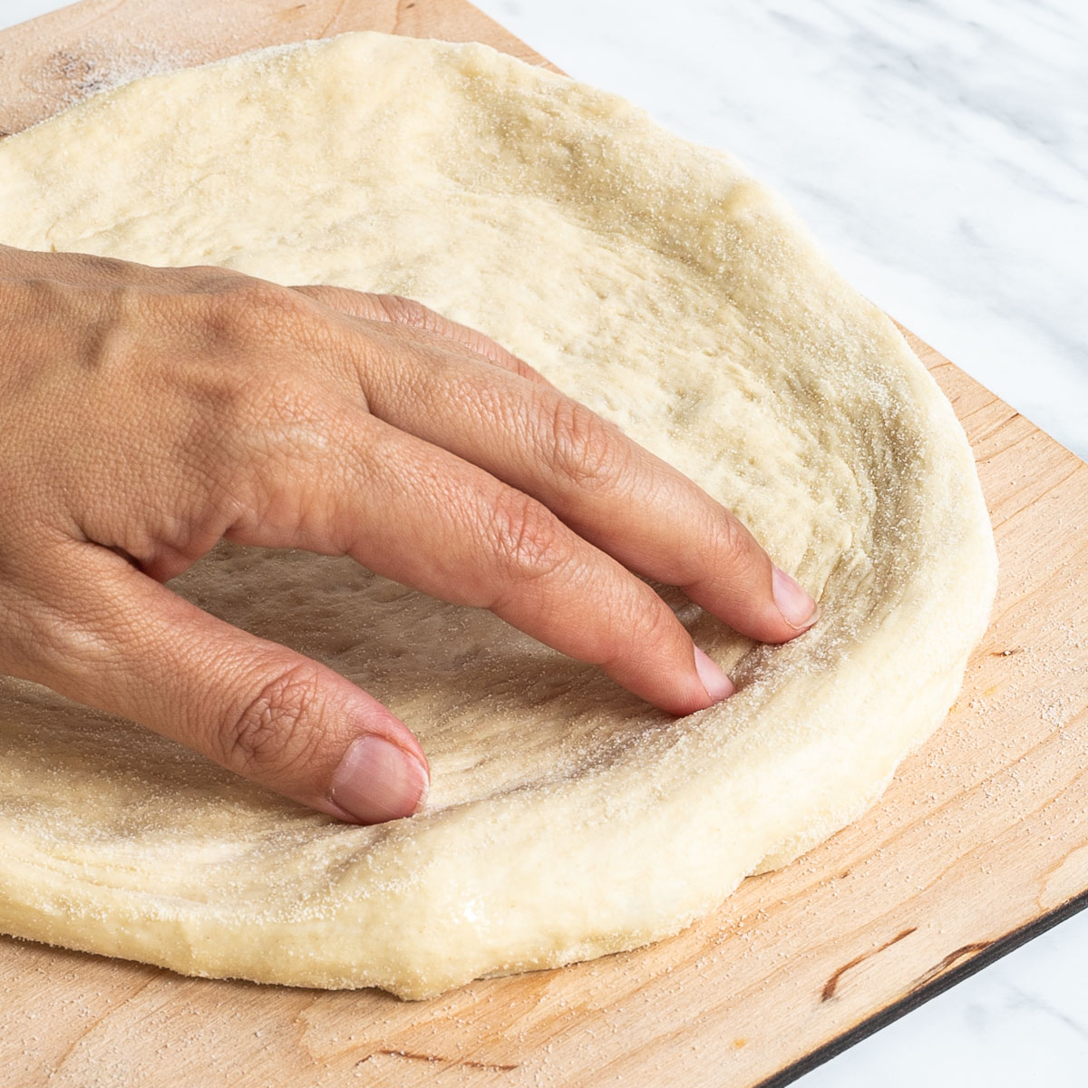 A hand is shaping the pizza dough on a lightly floured wooden pizza peel.