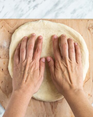 Two hands are shaping the pizza dough on a lightly floured wooden pizza peel.