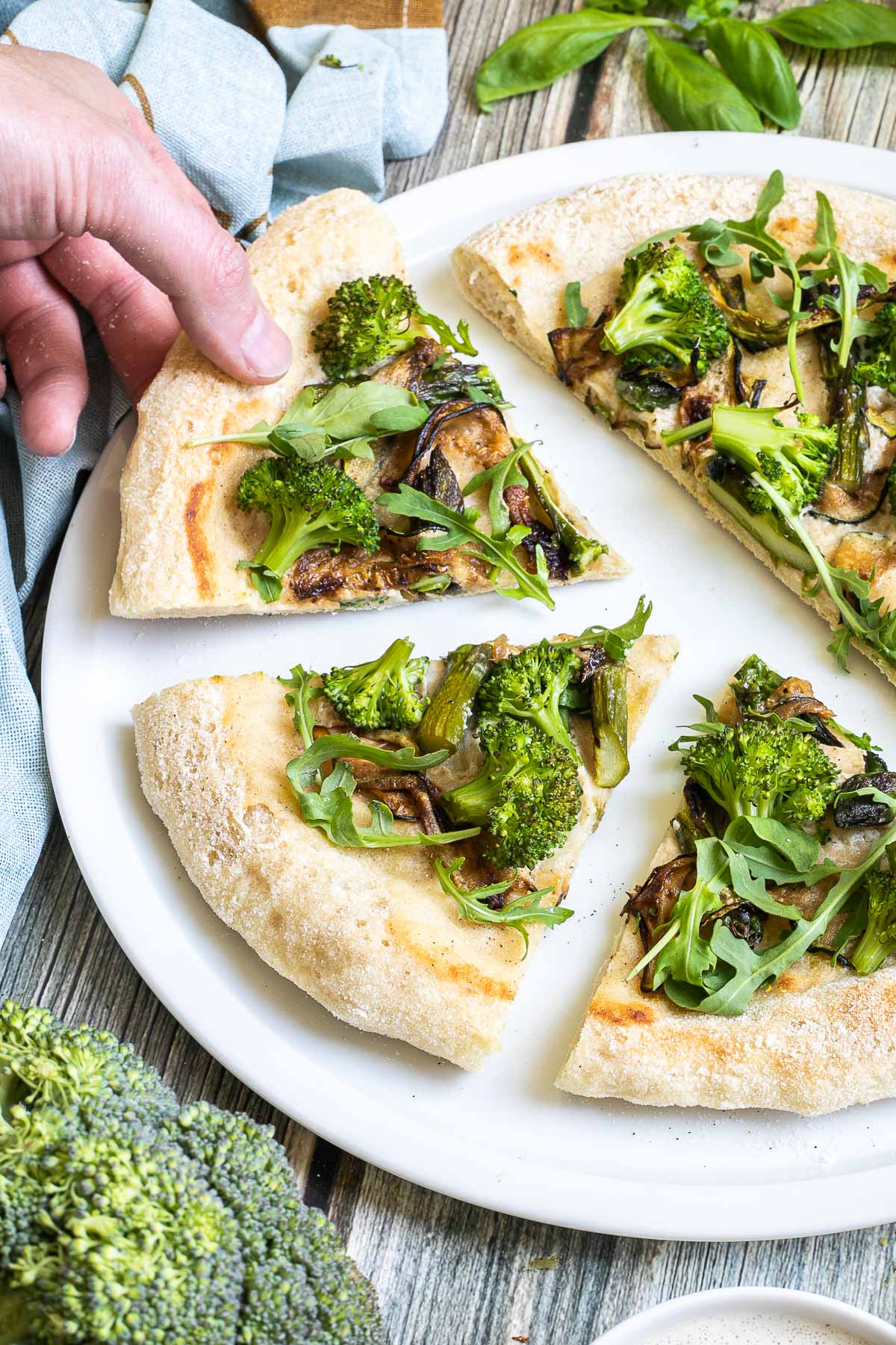 Sliced pizza on a white pizza plate topped with white sauce, zucchini, broccoli, asparagus, mushrooms, and fresh arugula. A hand is taking one slice.