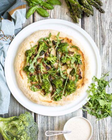 A pizza on a white pizza plate topped with white sauce, zucchini, broccoli, asparagus, mushrooms, and fresh arugula. More white sauce and fresh ingredients are scattered around the plate.