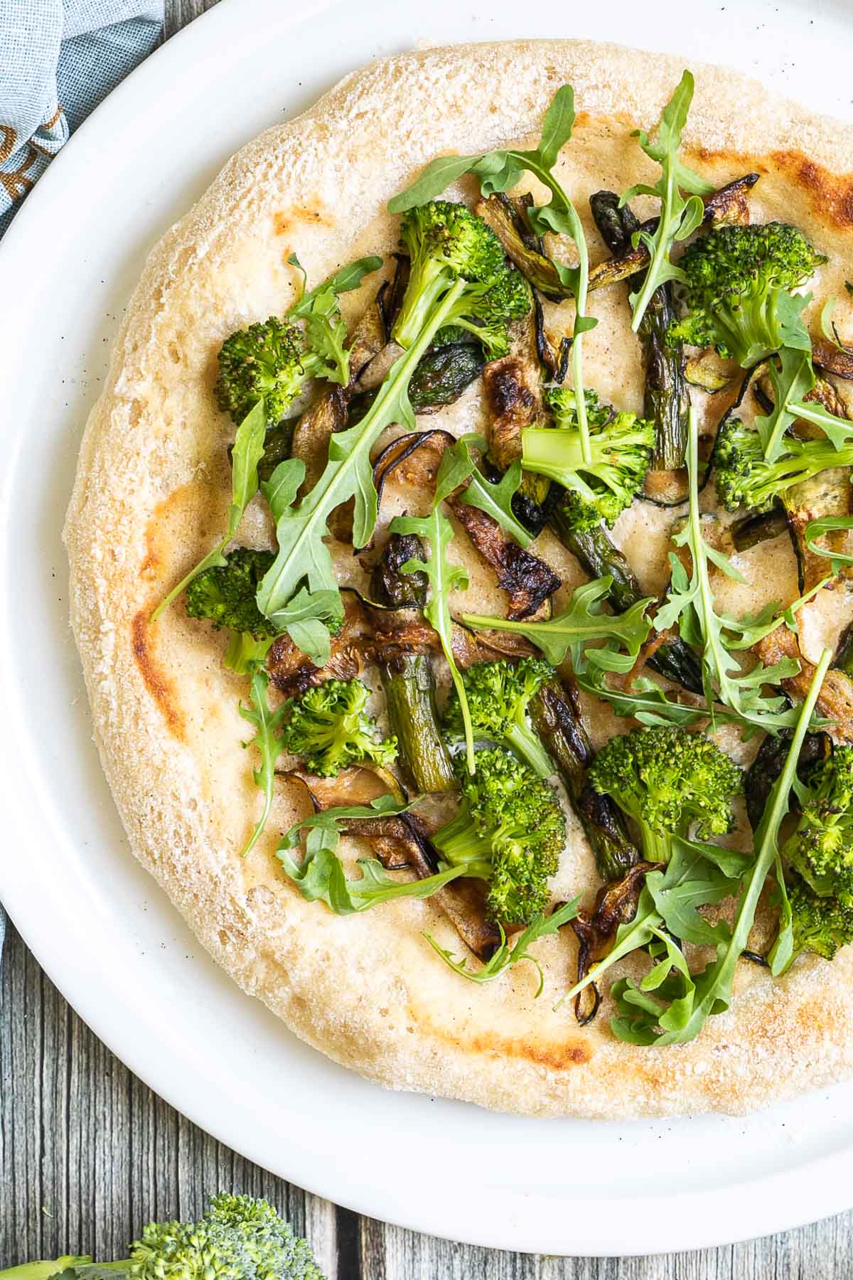 A pizza on a white pizza plate topped with white sauce, zucchini, broccoli, asparagus, mushrooms, and fresh arugula.