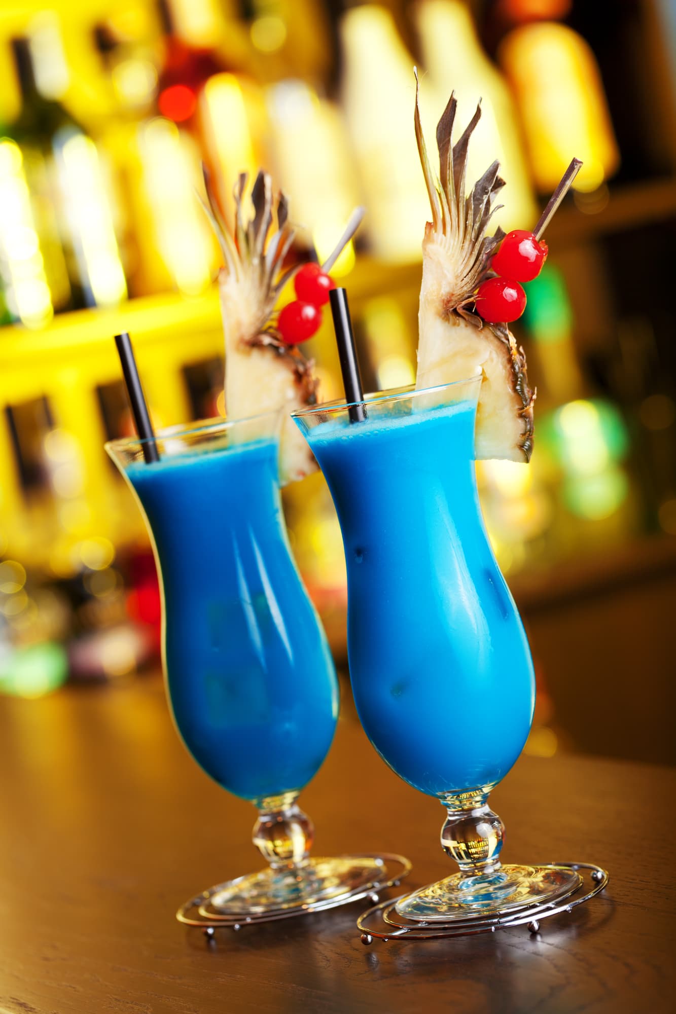 Creamy blue cocktail in two tall glasses, served with a small slice of pineapple and cherries.