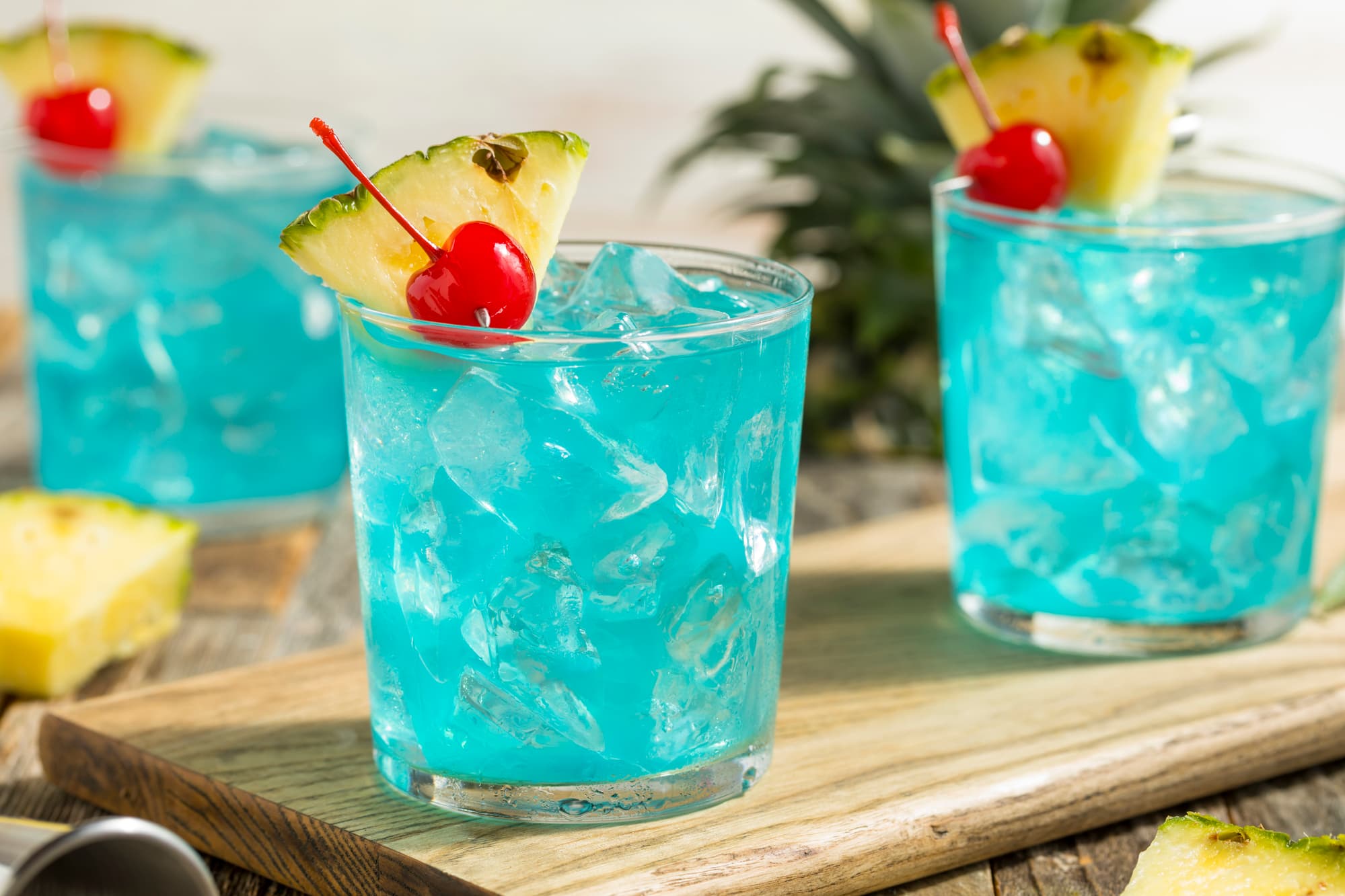 Light blue cocktail in two small glasses with lots of ice, served with a small slice of pineapple and cherries. 