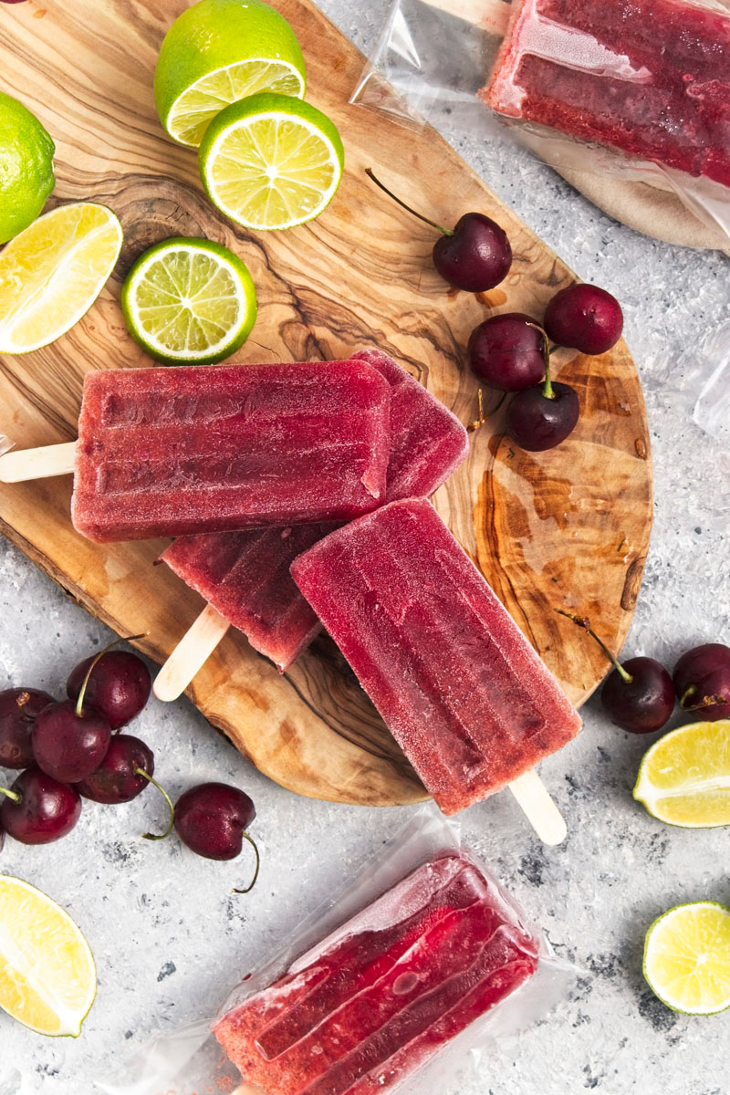 Vibrant purple popsicles on a wooden board. Cherries and lime slices are around them.