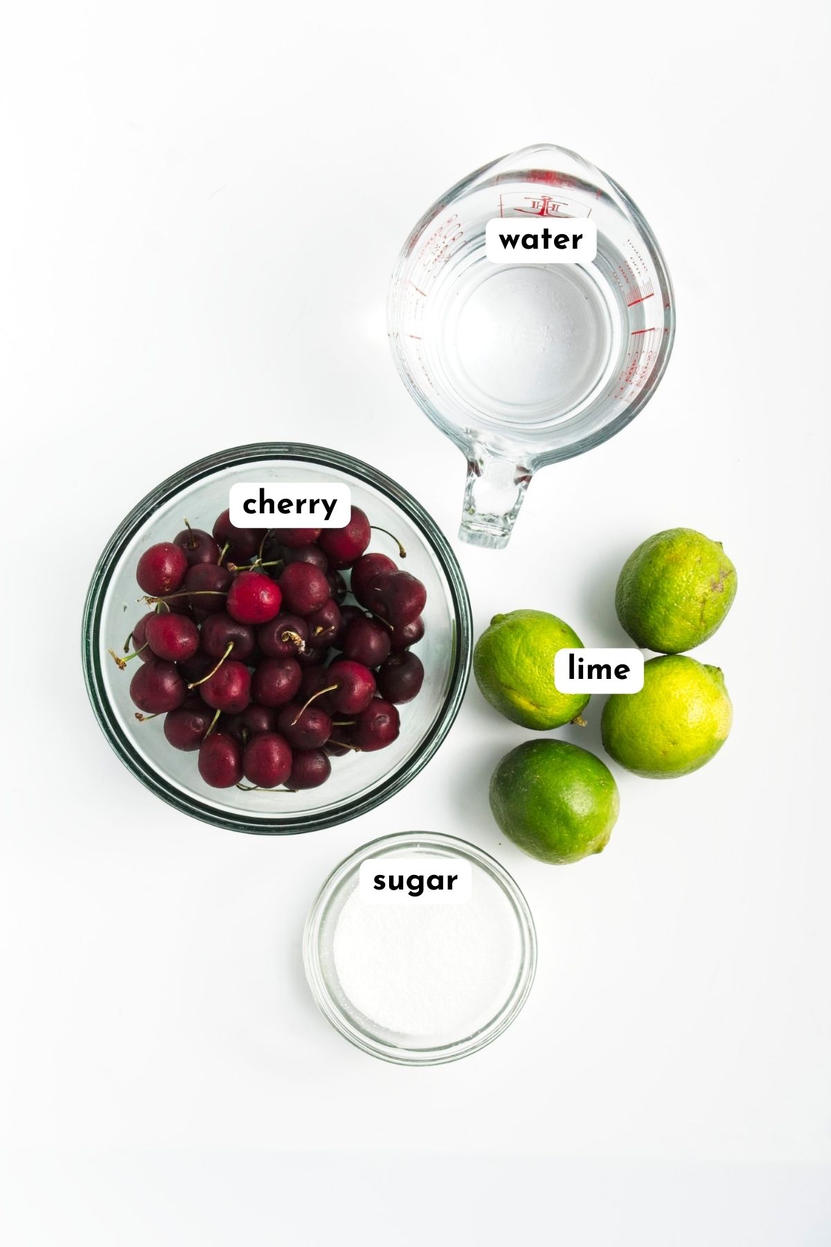 Ingredients of cherry popsicles on a white surface like jug of water, bowl of cherries, 4 limes, and a small bowl of white sugar
