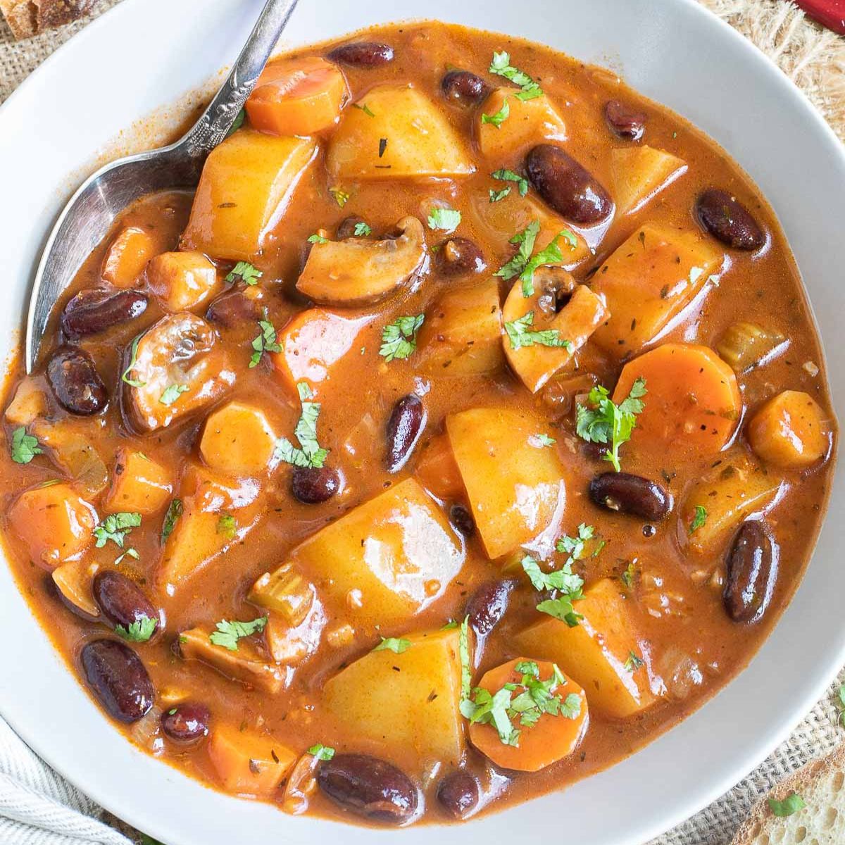 Vegan stew in a white bowl with lots of chopped vegetables like potatoes, carrots, mushroom, celery and red kidney beans. Topped with freshly chopped green herbs.