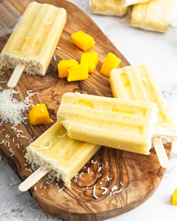 Vibrant yellow popsicles on a wooden board. Mango pieces and shredded coconut is around them.