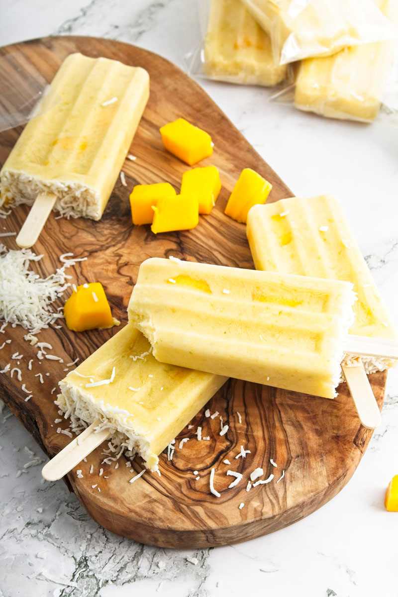 Vibrant yellow popsicles on a wooden board. Mango pieces and shredded coconut is around them. 