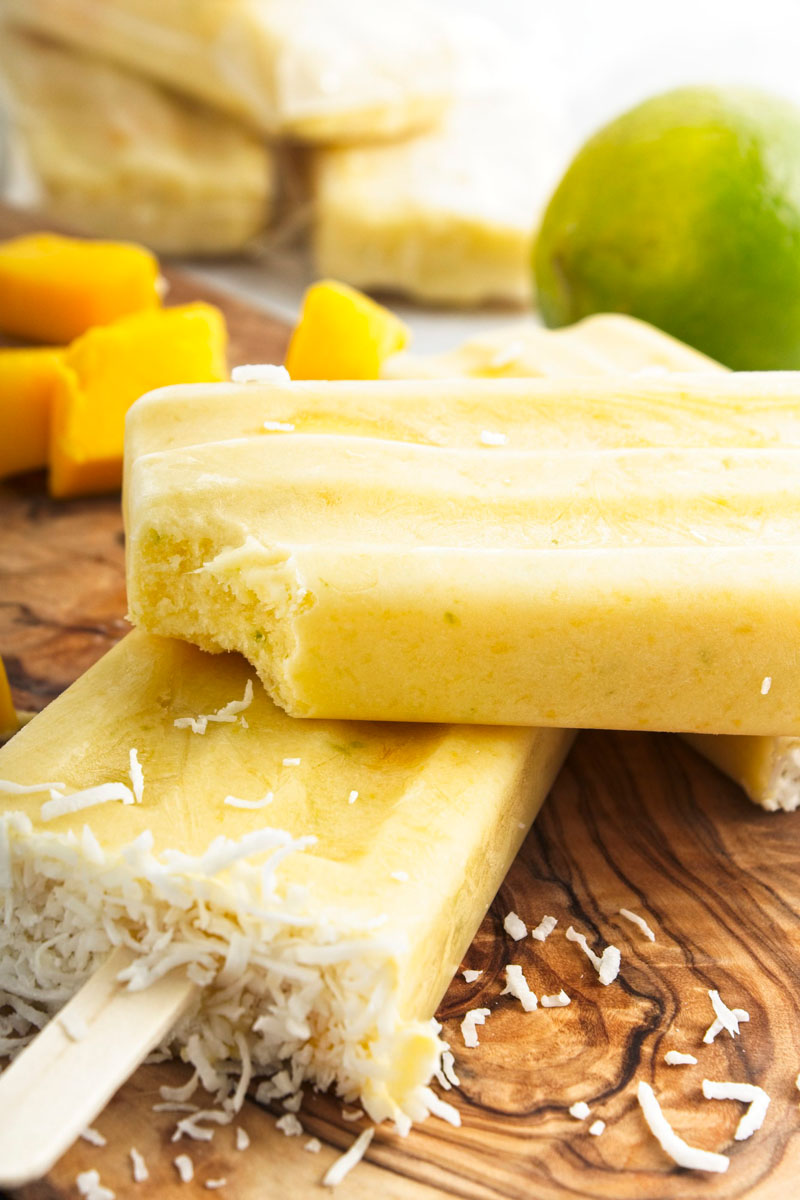 Vibrant yellow popsicles on a wooden board. Mango pieces and shredded coconut is around them. A bite is missing from one of them.