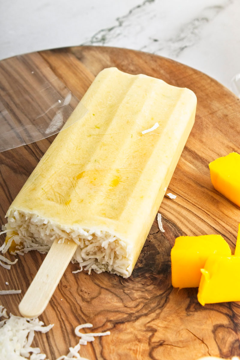 Vibrant yellow popsicles on a wooden board. Mango pieces and shredded coconut is around them.