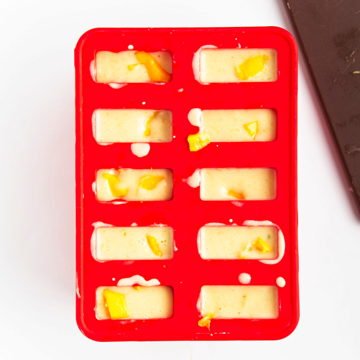 Red popsicle mold with 10 holes filled to the brim with yellow puree and mango pieces.