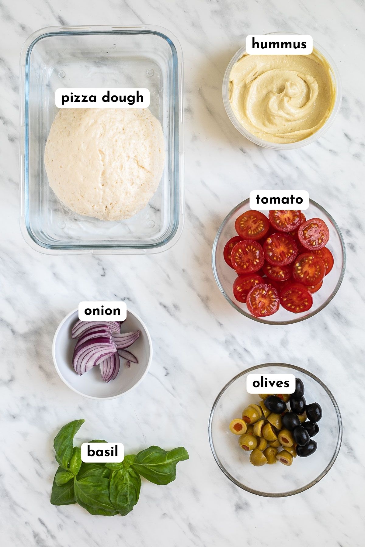 Ingredients of hummus pizza in small containers: pizza dough, hummus, cherry tomatoes, red onion, basil, and olives.