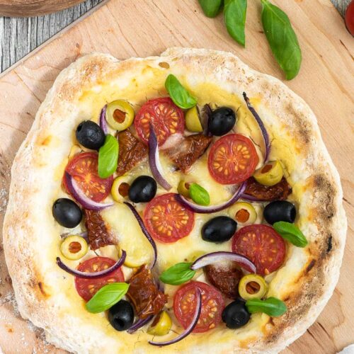Pizza on a wooden baker's peel topped with light brown hummus, cherry tomatoes, green and black olives, red onion slices, and fresh basil leaves. More hummus and other fresh ingredients are scattered around it.