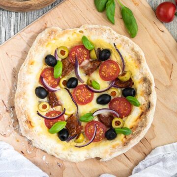 Pizza on a wooden baker's peel topped with light brown hummus, cherry tomatoes, green and black olives, red onion slices, and fresh basil leaves.