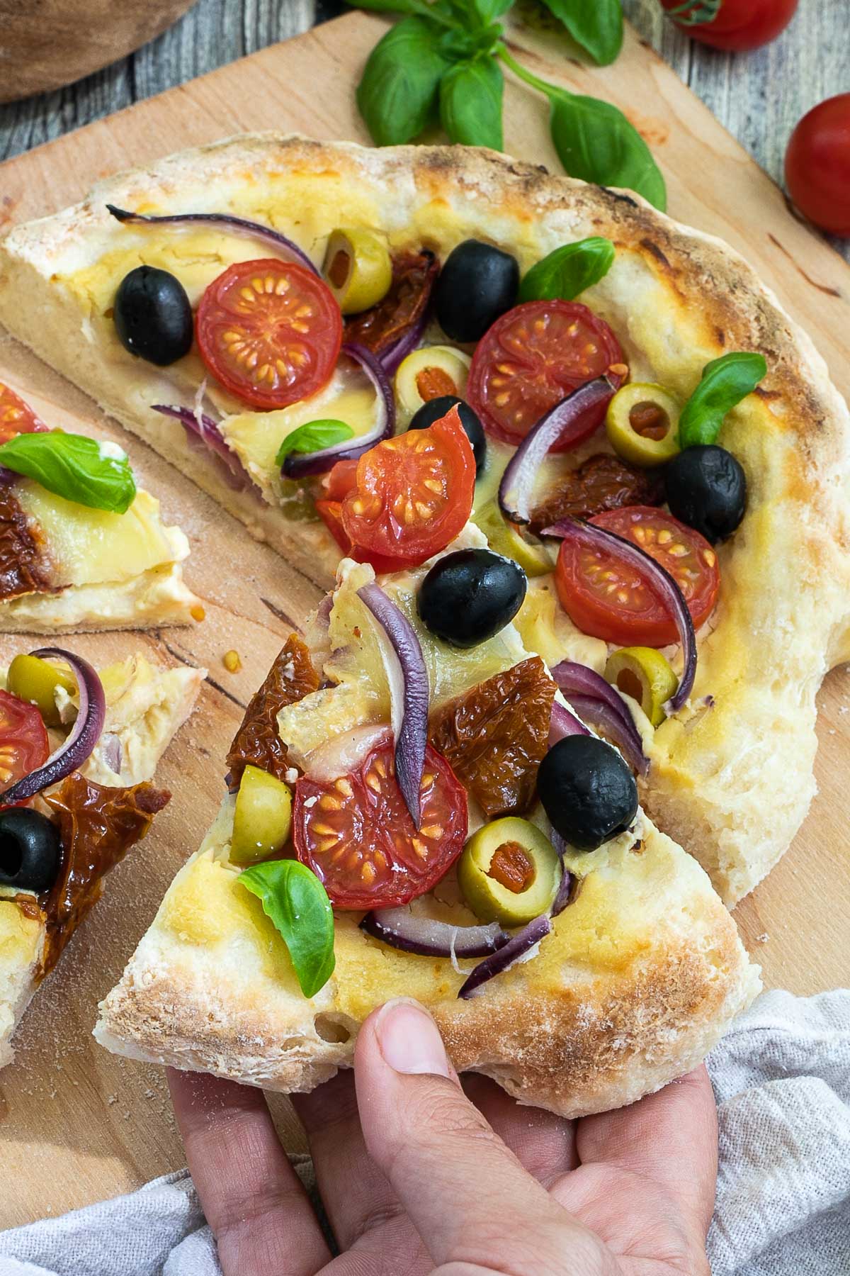 Sliced pizza on a wooden baker's peel topped with light brown hummus, cherry tomatoes, green and black olives, red onion slices, and fresh basil leaves. A hand is talking a slice.