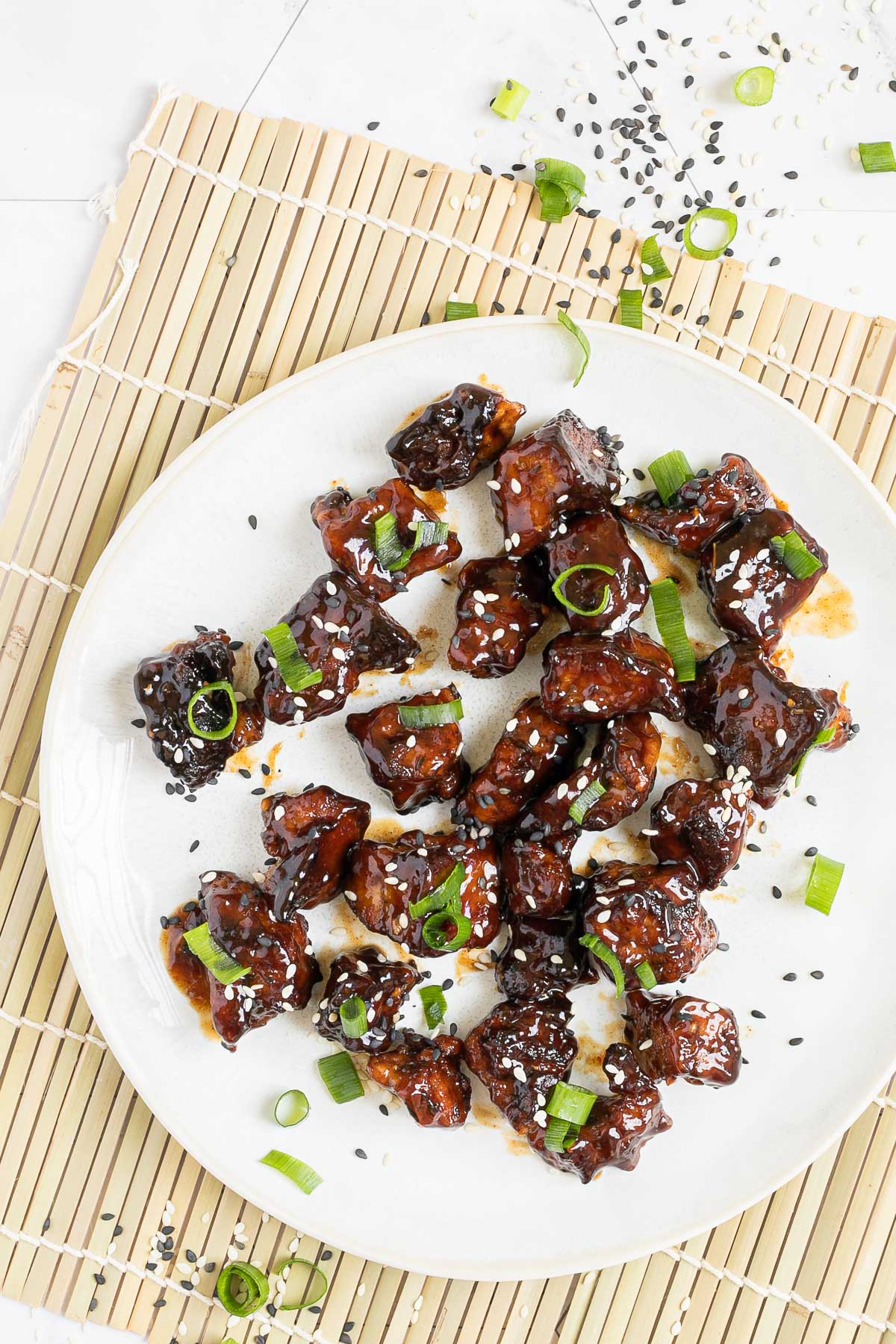 Dark brown sticky juice tofu pieces on a white plate sprinkled with black and white sesame seeds and chopped scallion.