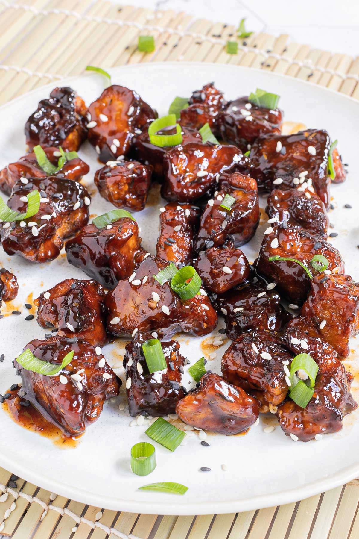 Dark brown sticky juice tofu pieces on a white plate sprinkled with black and white sesame seeds and chopped scallion.