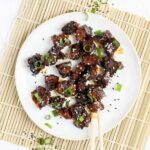 Dark brown sticky juice tofu pieces on a white plate sprinkled with black and white sesame seeds and chopped scallion. The end of a chopsticks is about to grab a piece.