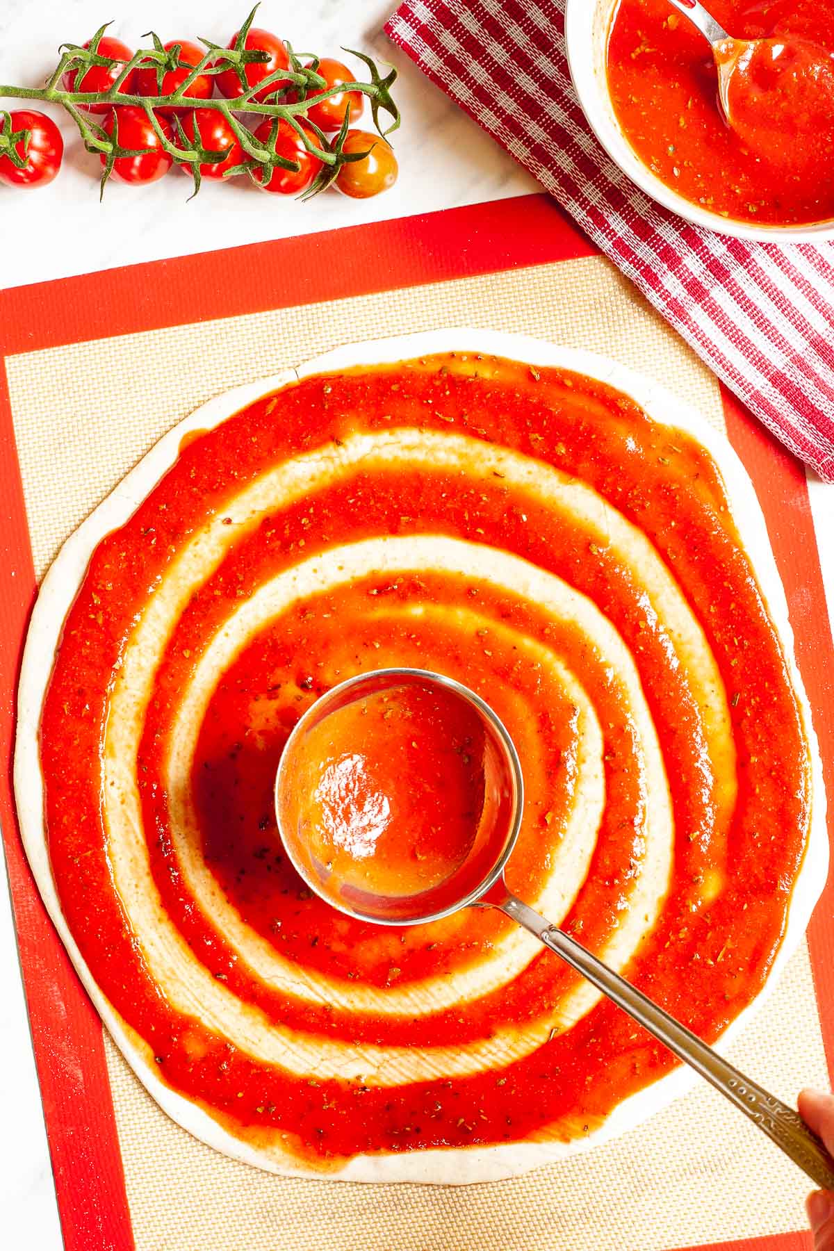 Red pizza sauce is spread in circles on top of a pizza crust with a ladle.