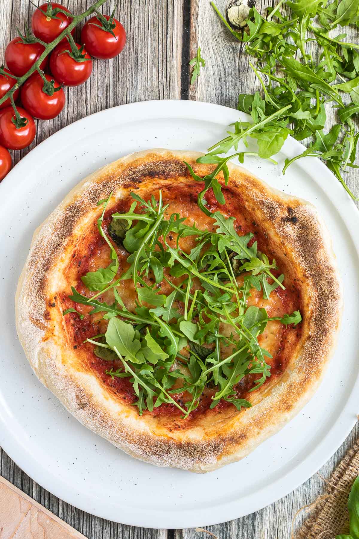 Sliced pizza topped with tomato sauce, melted cheese and fresh arugula on a white pizza plate. Fresh ingredients are around it.
