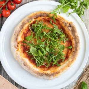 Sliced pizza topped with tomato sauce, melted cheese and fresh arugula on a white pizza plate. Fresh ingredients are around it.