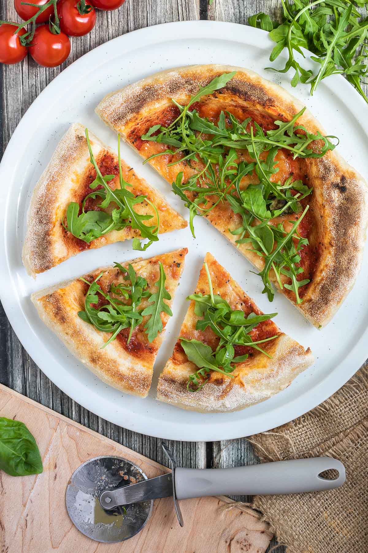 Sliced pizza topped with tomato sauce, melted cheese and fresh arugula on a white pizza plate. Fresh ingredients are around it.