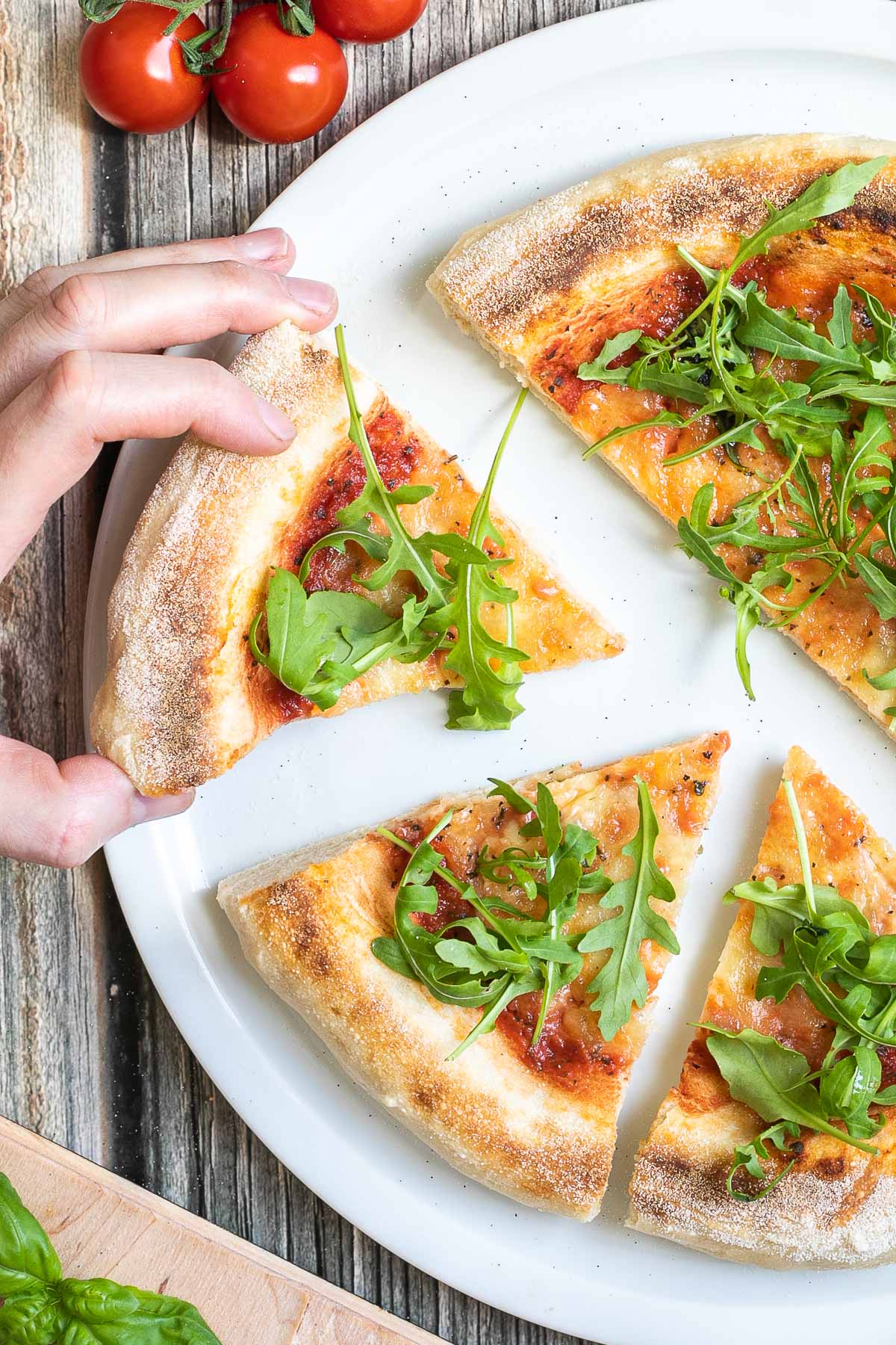 A pizza cut into slices with fluffy crispy crust topped with tomato sauce, melted cheese and fresh arugula on a white plate. A hand is taking one slice.