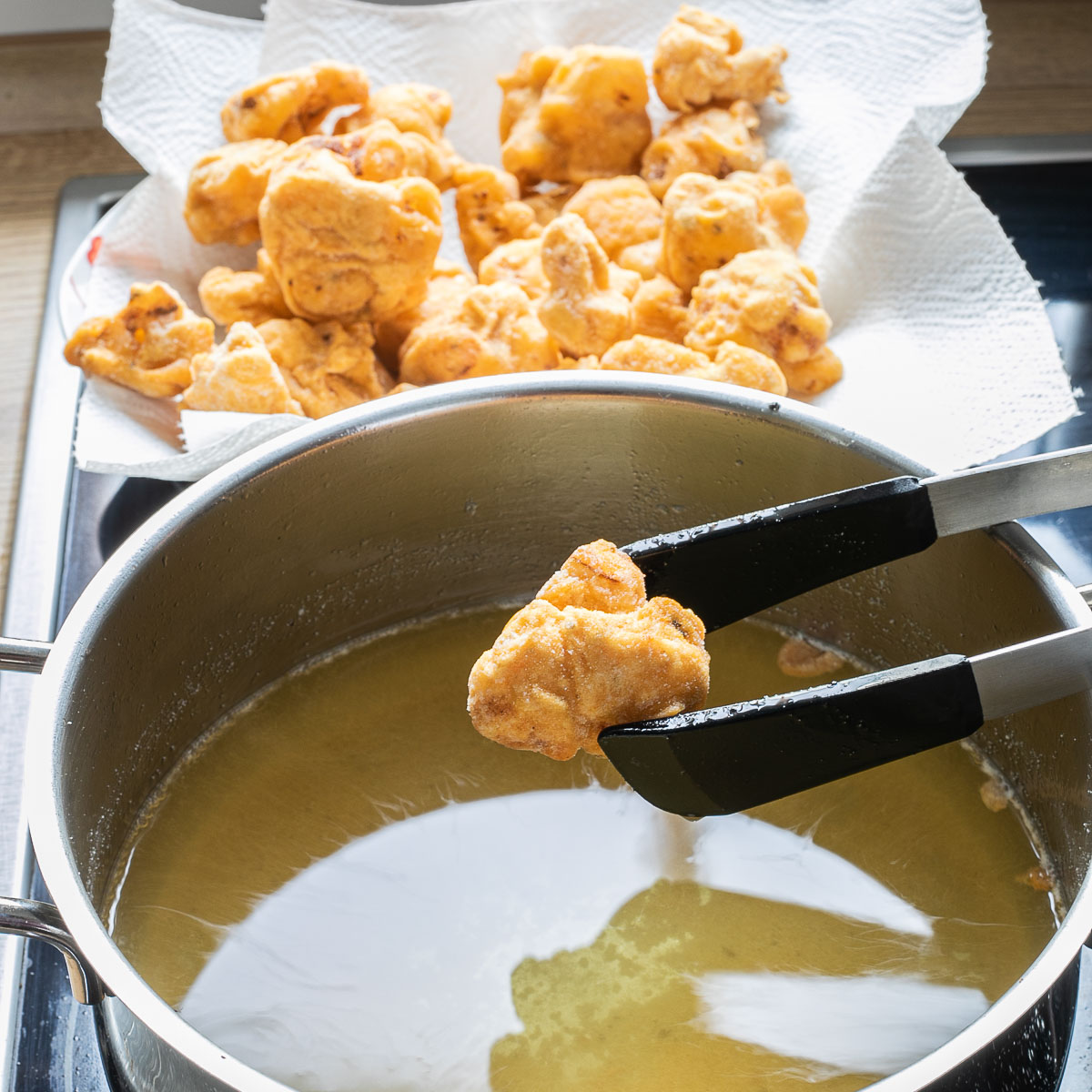 A large pot with oil. Tongs are holding one breaded and crispy cauliflower floret. Other florets are ready and behind the pot on white paper towel.