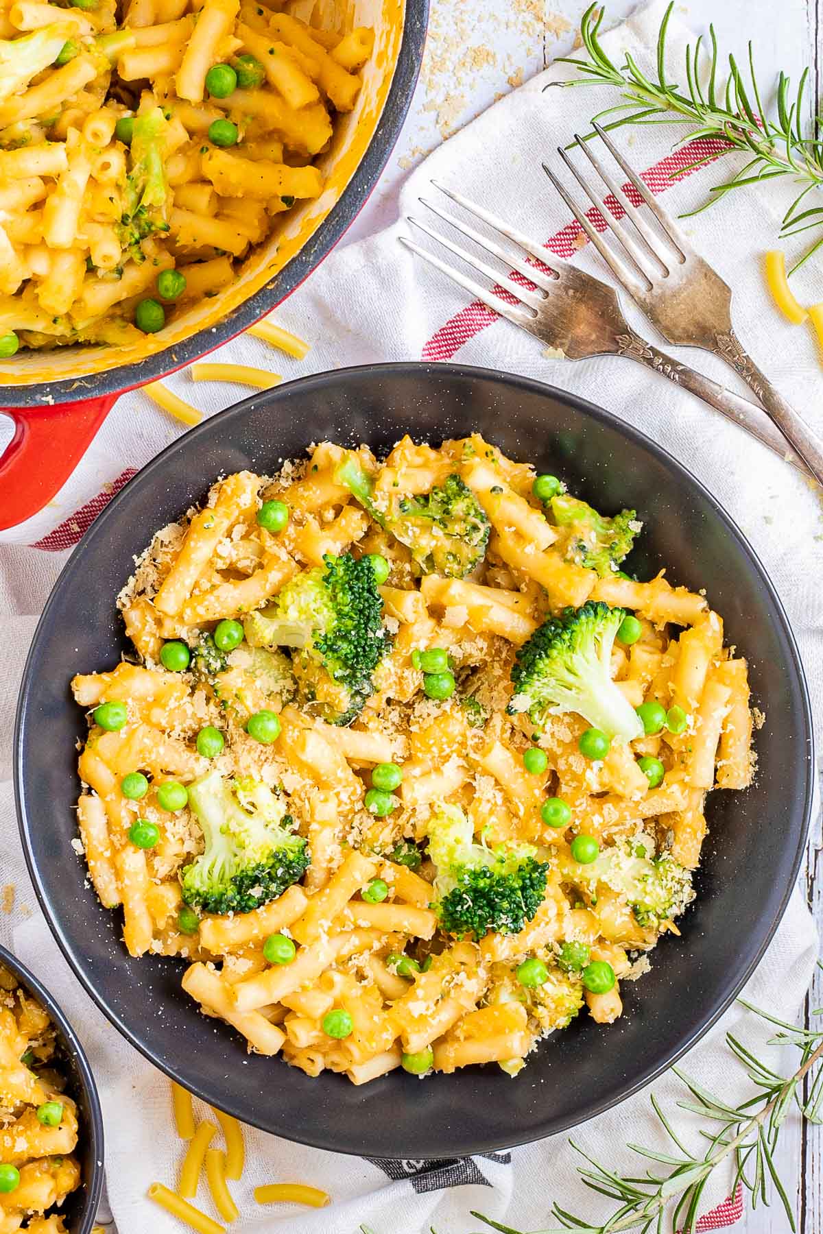 A red white enameled Dutch oven and 2 black bowls are serving short pasta in thick yellow sauce with broccoli florets and green peas added as toppings. 