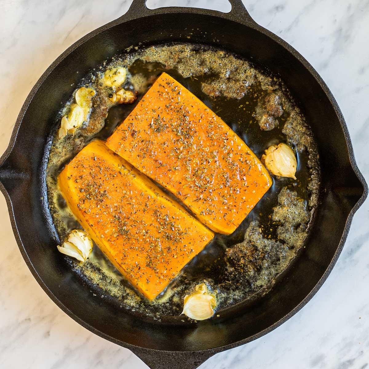 Cast iron skillet with two large squash slices and some garlic cloves in bubbling melted butter. 