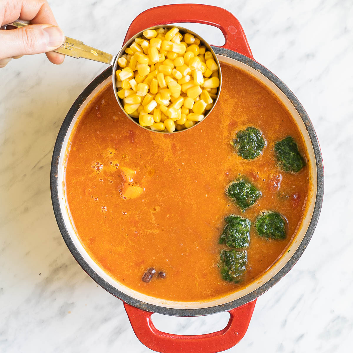 Orange soup in a white-red enameled Dutch oven with green frozen spinach cubes. A hand is holding a cup to add sweet corn.