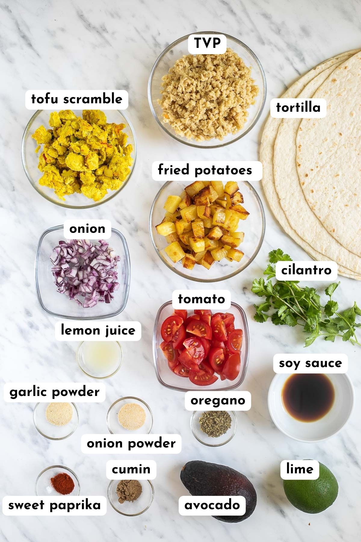 Ingredients of breakfast enchiladas are in small glass bowls like tofu scramble, tvp, diced potatoes, tomatoes, onion, cilantro, avocado, lime and many spices and herbs. 