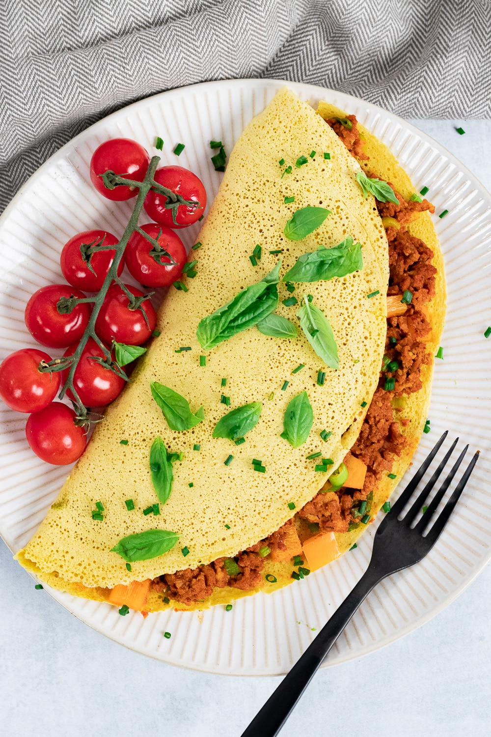 Yellow omelette folded in half stuffed with brown crumbles sprinkled with freshly cut herbs and cherry tomatoes on a vine.