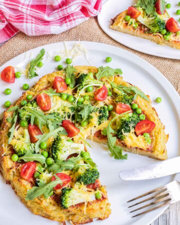 Hash brown crust pizza with topped with tomato sauce, broccoli, peas cherry tomatoes, arugula, and shredded melted cheese. Served on a white plate. A slice is missing and a fork and knife are placed instead there.