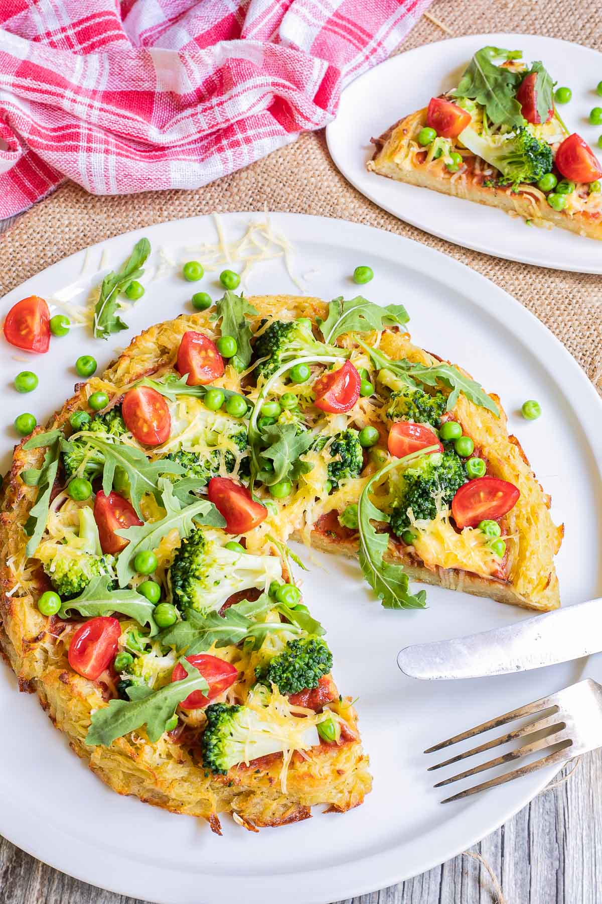 Hash brown crust pizza with topped with tomato sauce, broccoli, peas cherry tomatoes, arugula, and shredded melted cheese. Served on a white plate. A slice is missing and a fork and knife are placed instead there.